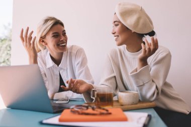 Neşeli Asyalı ve Kafkasyalı en iyi arkadaşlar kafenin içindeki olumlu toplantı sırasında web yayınını tartışıyor, modern dizüstü bilgisayarlı mutlu blog yazarları içeride gülüyor ve eğleniyorlar.