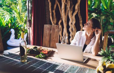Dizüstü bilgisayarında web makalesi yaratırken içerik yayını için bir bardak beyaz şarap düşünen düşünceli kadın serbest yazar, netbook üzerine düşünen düşünceli grafik tasarımcısı