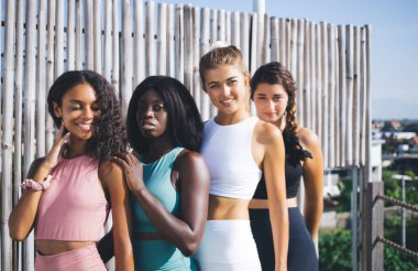 Aktif durumda olan çok ırklı kızlar açık havada yoga seansı için kameraya bakıyorlar. Kilo kaybı ve vücut bakımı konsepti, spor kıyafetleri giymiş farklı kızların yarım boy portresi.