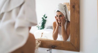 Kafasında havlu olan genç ve güzel bir bayan evdeki banyoda aynanın yansımasına gülümsüyor. 30 yaşında mutlu bir kadın günlük sabah ayinleri ve temizlik yapıyor. Sağlıklı cilt bakımı.