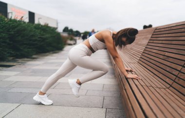 Spor kıyafetleriyle kenarda duran, ahşap bankta yaslanan ve şehir sokaklarında antrenman yaparken bacaklarını esneten kadın sporcuların yan görüntüsü.