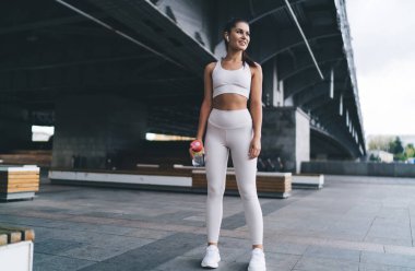 Tam boy kendine güvenen, elinde spor kıyafetleri ve kablosuz kulaklıklarla elinde su şişesi olan sporcu bir bayan sporcu. Sokakta spor yaparken gülümseyen ve gözlerini kaçıran.