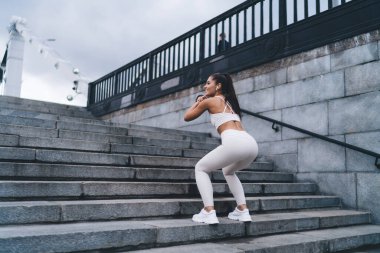 Genç bir kadın, kentteki sabah egzersizleri sırasında kalça kaslarını şişirmek ve formda, basit ve etkili egzersizler yapmak için çömelir. Sağlıklı yaşam tarzı, vücudunu sportif formda tutan bir kadın.