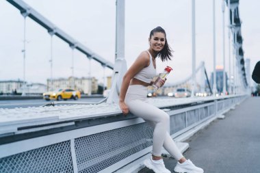 Spor kıyafetli atletik bir kadın köprüde oturmuş kameraya bakarken ve şehirde spor yaparken mola verirken görüntüsü.