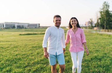 Parktaki çimenlerde yürürken el ele tutuşup kameraya bakan çok ırklı, pozitif genç bir çift.