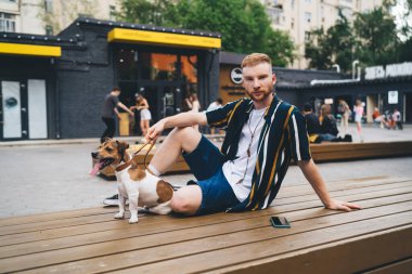 Günlük giysiler içindeki genç adam ahşap bankta oturuyor ve köpek tasması tutuyor modern parkın karşısında yürüyen insanlarla kameraya bakıyor.