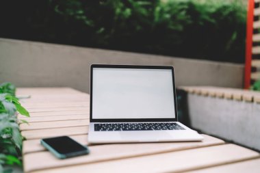 Boş ekranlı modern dizüstü bilgisayar şehirdeki uzaktan çalışma sırasında yeşil santrallere karşı cep telefonuyla ahşap masaya yerleştirilmiş.