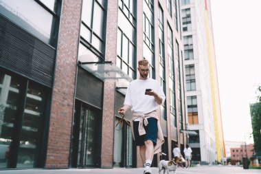 Gündüz vakti modern binaların yanında köpekle yürürken cep telefonundan mesaj atan rahat giyimli genç bir adam.