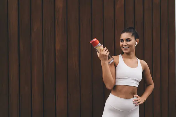 stock image Young fit female athlete in sportswear with bottle of lemon water listening to music using earbuds and smiling while standing and looking away after workout against wooden wall