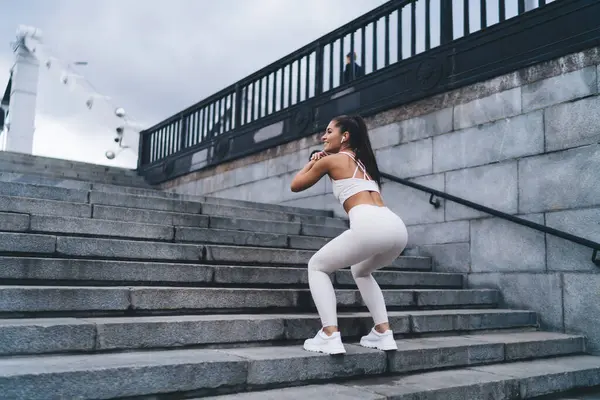 stock image Young woman makes squats to pump the muscles of buttocks and make fit form, simple and effective exercises during morning workout in city. Healthy lifestyle, female keeping the body in sporty shape