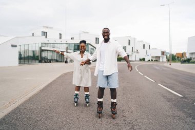 Keyifli Afro-Amerikan erkek ve kadın günlük kıyafetleriyle tekerlekli sandalyelere biniyor ve şehir evlerinin yakınındaki asfalt yolda el ele tutuşuyorlar.