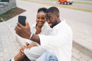 Mutlu etnik bir adam şehirde merdivenlerde oturmuş eğlenirken akıllı telefondan gülümseyen bir kadınla selfie çekiyor.