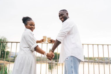 Genç, Afro-Amerikalı kadın ve erkeğin el ele tutuşup kağıt bardaklarda sıcak içecekler içerken ve göllü şehir parkında omzunun üzerinden kameraya bakarken.
