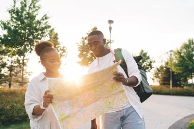 Günbatımında yeşil ağaçlar ve çalılarla parktaki kaldırımlı yolda duran ve haritayı inceleyen gündelik giysili genç siyah sevgili ve kız arkadaş.