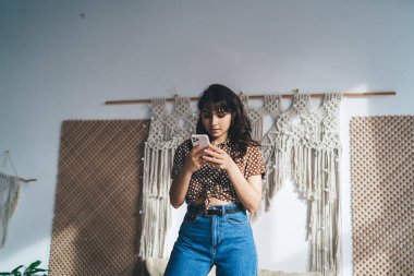 Puantiyeli bluzlu, ciddi bir genç kadın. Yatağın üzerinde duruyor ve duvarın yanındaki cep telefonuyla internette sörf yapıyor. Makrome süsleme ve rattan süslemelerle süslenmiş.