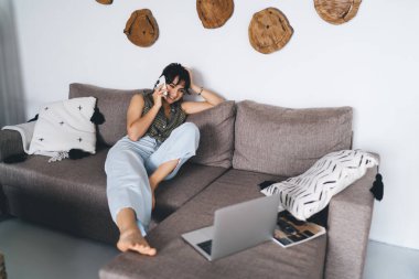 Oturma odasında ahşap süslemelerle konuşurken rahat koltukta dinlenen ve dizüstü bilgisayarla ilginç filmler izleyen gülümseyen genç bir kadın.