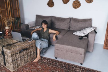 Yüksek açılı, serbest çalışan, gülümseyen ve bilgisayarla koltuğa oturan genç bir kadın uzaktan kumanda ile evden uzaklaşıyor.