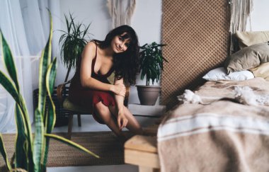 Kırmızı elbiseler giyen, evdeki rahat koltukta oturan ve gülümseyerek kameraya bakan genç ve şehvetli bir kadın.