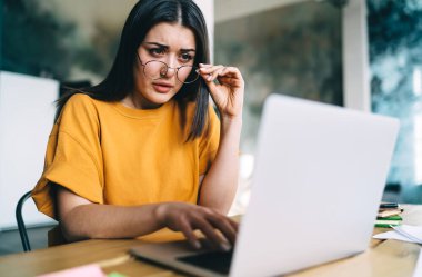 Turuncu tişörtlü, masa başında oturan ve iş sırasında ağzı açık bir şekilde ekrana bakarken laptop kullanan şüpheli bir kadın.