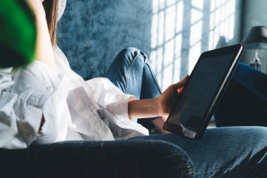 Kanepede oturan ve güneşli oturma odasında tablet kullanan sıradan giyinmiş, isimsiz bir kadının yan görüntüsü.