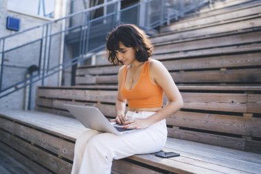 Gülümseyen, kısa kıvırcık saçlı, sıradan elbiseli, ahşap merdivenlerde akıllı telefonun yanında oturan ve uzaktan bilgisayarla çalışan serbest çalışan genç bir bayan.