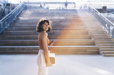 Gülümseyen, serbest çalışan, günlük kıyafetler giyen, ahşap merdivenlerin yanında duran, akıllı telefonlara bakan, şehirdeki kameralara bakan genç bir kadın.
