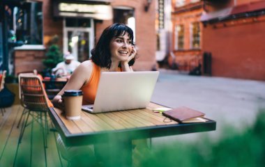 Genç bayan dijital göçebe ahşap masada oturuyor ve dizüstü bilgisayarla çalışırken eline yaslanıyor ve sokak kafesinde gülümsüyor.