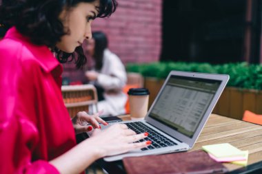 Kafe terasından uzaktan çalışırken ahşap masada dizüstü bilgisayarla oturan pembe bluzlu genç, konsantre kadın serbest çalışanın yan görüntüsü