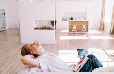 Orta yaşlı kadın, günlük kıyafetlerle, dergilerle kanepede uzanmış, elleri başının arkasında dizüstü bilgisayar kullanarak evden uzakta çalışırken yan görüş.