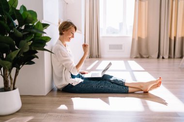 Orta yaşlı heyecanlı kadın serbest çalışanın yumruğunu sıkması ve evden iş sırasında dizüstü bilgisayar ekranına bakarken başarıyı kutlamasının yan görüntüsü