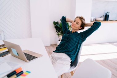 Masada dizüstü bilgisayarla oturmuş gülümseyen yetişkin bir kadın. Düşünceli bir şekilde ekranı seyrediyor ve evdeki işi bitirdikten sonra dinleniyor.