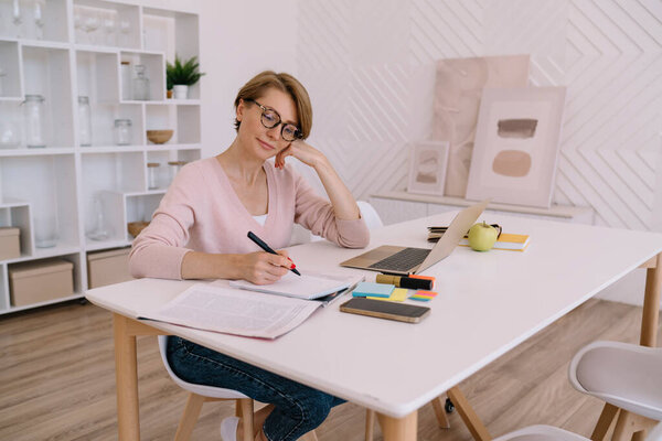 Smiling adult woman in casual clothes and eyeglasses sitting at white table with smartphone and laptop and taking notes while doing freelance job in light modern room