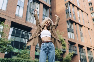 Düşük açılı gülümseyen siyah kadın gündelik giysiler içinde kulaklıkla müzik dinliyor kolları havada ve hafta sonu sokakta dans ediyor.