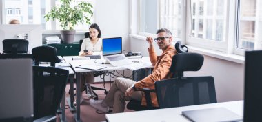 Gündelik giysiler içindeki pozitif iş adamı laptopla masada oturuyor ve modern ofiste çalışırken kadın iş arkadaşının yanından uzaklaşıyor.