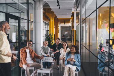 Çağdaş çalışma ortamında cam duvarın yanında oturan çeşitli iş adamları iş stratejisi hakkında konuşan meslektaşlarına bakıyorlar.