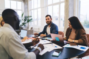 Farklı erkek ve kadın meslektaşları olan sakallı bir adam yeni iş stratejilerini tartışırken masalarında aletler ve kağıtlarla bir araya geliyorlar.