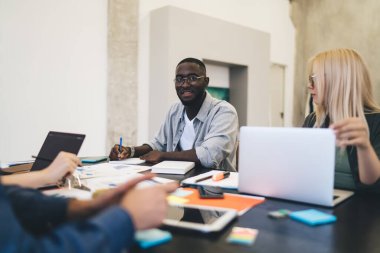 Afrika kökenli Amerikalı iş adamı çeşitli meslektaşlarıyla masaya otururken modern ofiste proje üzerinde çalışırken notlar yazıyor.