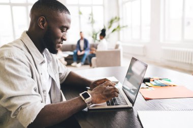 İş yerinde çalışırken, masa başında oturup dizüstü bilgisayarda yazan, günlük kıyafetlerle çalışan Afrikalı Amerikalı erkek işçilere yandan bakıldığında,