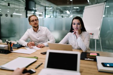 Resmi kıyafetli erkek ve kadın iş arkadaşları ofiste masada oturuyor ve toplantı sırasında mahsul arkadaşını dikkatle dinliyorlar.