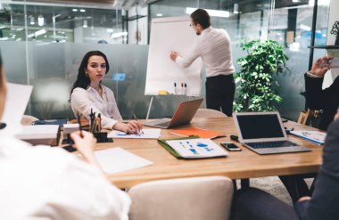 Resmi giysiler içinde bir grup genç iş adamı dizüstü bilgisayarlarla ve belgelerle masada oturuyor ve beyin fırtınası sırasında modern ofiste birlikte çalışıyorlar.