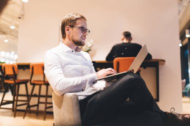 Gözlüklü, resmi beyaz gömlekli, dizüstü bilgisayarlı, projede çalışırken ekrana bakan ve modern ofiste bacak bacak bacak üstüne atmış sakallı yöneticinin yan görüntüsü.