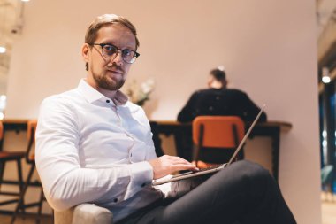 Gözlüklü ve resmi tişörtlü, modern ofisteki dizüstü bilgisayarla proje üzerinde çalışan ve kameraya bakan konsantre iş adamlarının düşük açılı görüntüsü.