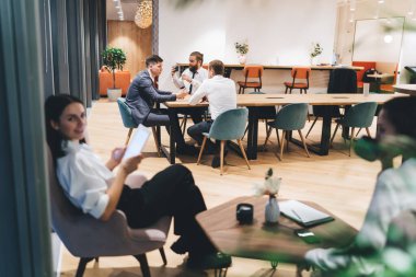 Resmi kıyafetler giymiş bir grup iş arkadaşı ahşap masalarda oturup geniş ofis kafeteryasında kahve içerken iş fikirlerini tartışıyorlar.