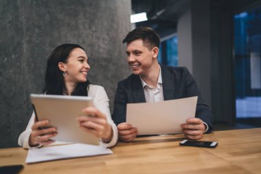 Resmi giysiler içinde gülümseyen adam ve kadın tabletle ahşap masada oturuyor ve modern ofiste birbirlerine bakarken proje detaylarını tartışıyorlar.