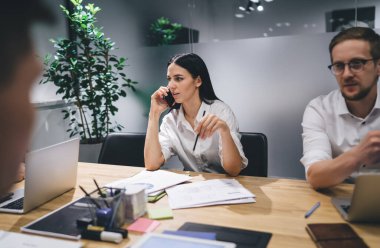 Ciddi bir iş kadını, konferans masasında meslektaşlarıyla oturup modern ofis yönetim kurulunda iş projesini tartışırken akıllı telefondan konuşuyor.