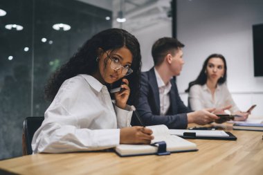 Ciddi Afrikalı Amerikalı iş kadını, toplantı sırasında toplantı masasında iş arkadaşlarıyla otururken akıllı telefondan konuşuyor ve organizatörle yazıyor.