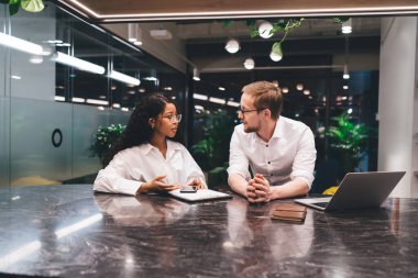 Ciddi erkek ve siyahi iş arkadaşları resmi giysiler içinde ellerinde aletlerle oturmuş modern ofiste iş projelerini tartışıyorlar.