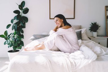 Gece elbiseli gülümseyen genç bayanın yan görüntüsü. Hafta sonu modern yatak odasında yatağında dinleniyor ve başka tarafa bakıyor.