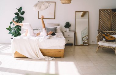 Neşeli çıplak ayaklı kadın, yatak odasında güneşli bir sabahta boş vakitte oynarken rahat bir yatakta uzanıyor ve ellerle yastığı havaya fırlatıyor.