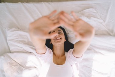 Mutlu kadın manzarası, gözleri kapalı rahat bir yatakta sırt üstü uzanıyor ve evde boş zaman geçirirken ellerini kaldırıyor.